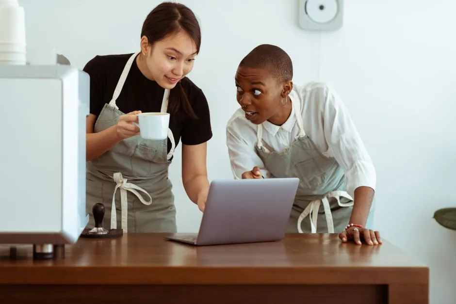 Cheerful surprised diverse colleagues in aprons using modern netbook and sharing business ideas while gathering at table with cup of drink in light cozy kitchen