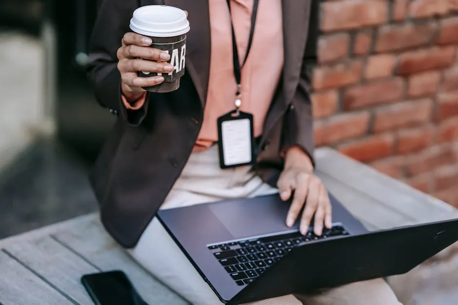Crop anonymous female entrepreneur wearing elegant black jacket with badge drinking coffee while using laptop sitting on bench